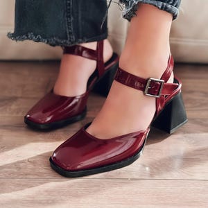 May include: Burgundy patent leather heeled sandals with a square toe and ankle strap. The sandals have a black block heel and a silver buckle closure. The shoes are on a wooden floor.