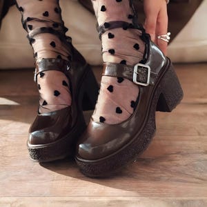 Può includere: Scarpe Mary Jane marroni con plateau e chiusura con fibbia. Le scarpe hanno un tacco grosso e una suola strutturata. Le scarpe in vernice sono indossate con collant neri a fantasia di cuori.