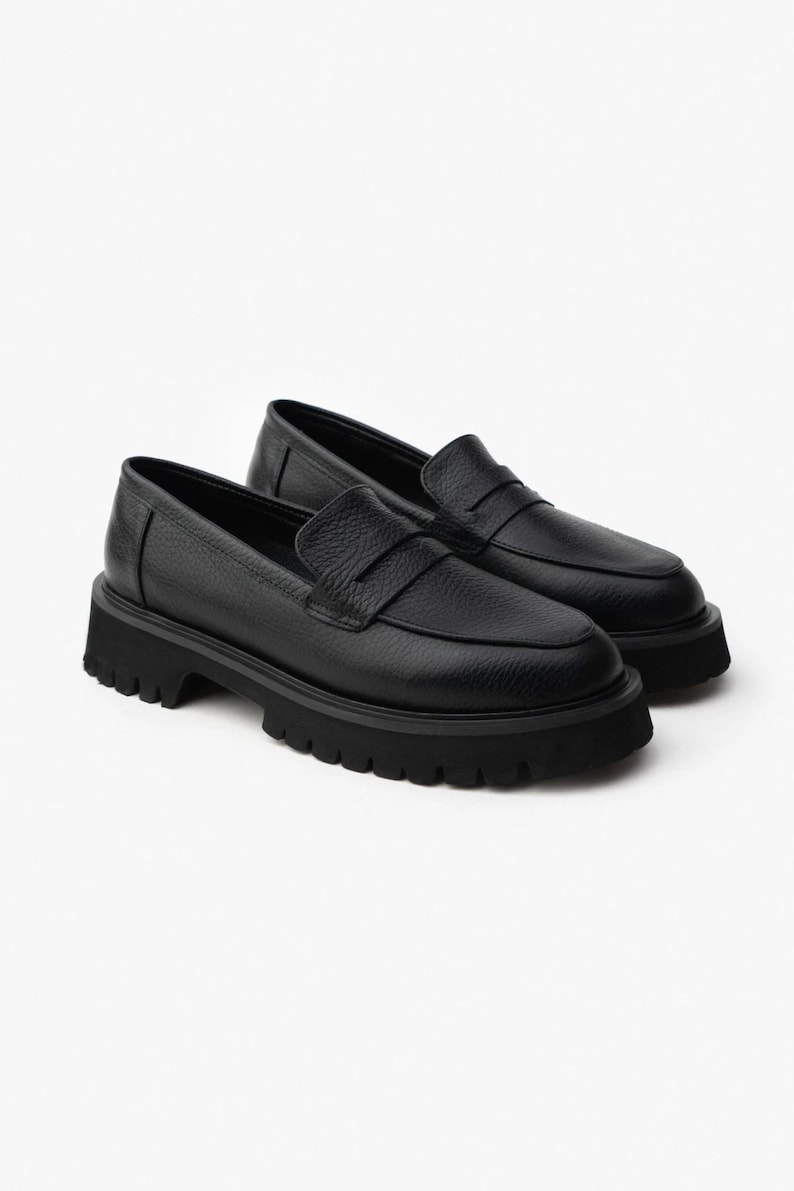 May include: A pair of black leather loafers with a chunky, lugged sole. The shoes have a classic penny loafer design with a rounded toe and a textured leather upper. The soles are thick and provide a rugged look.