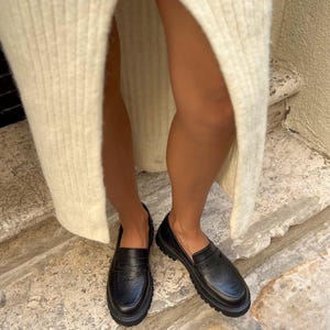 May include: Black leather loafers with a textured finish. The shoes have a chunky sole and a classic penny loafer design. The image shows the shoes from a low angle, highlighting their shape and texture. The loafers are worn with a cream-colored knit skirt.