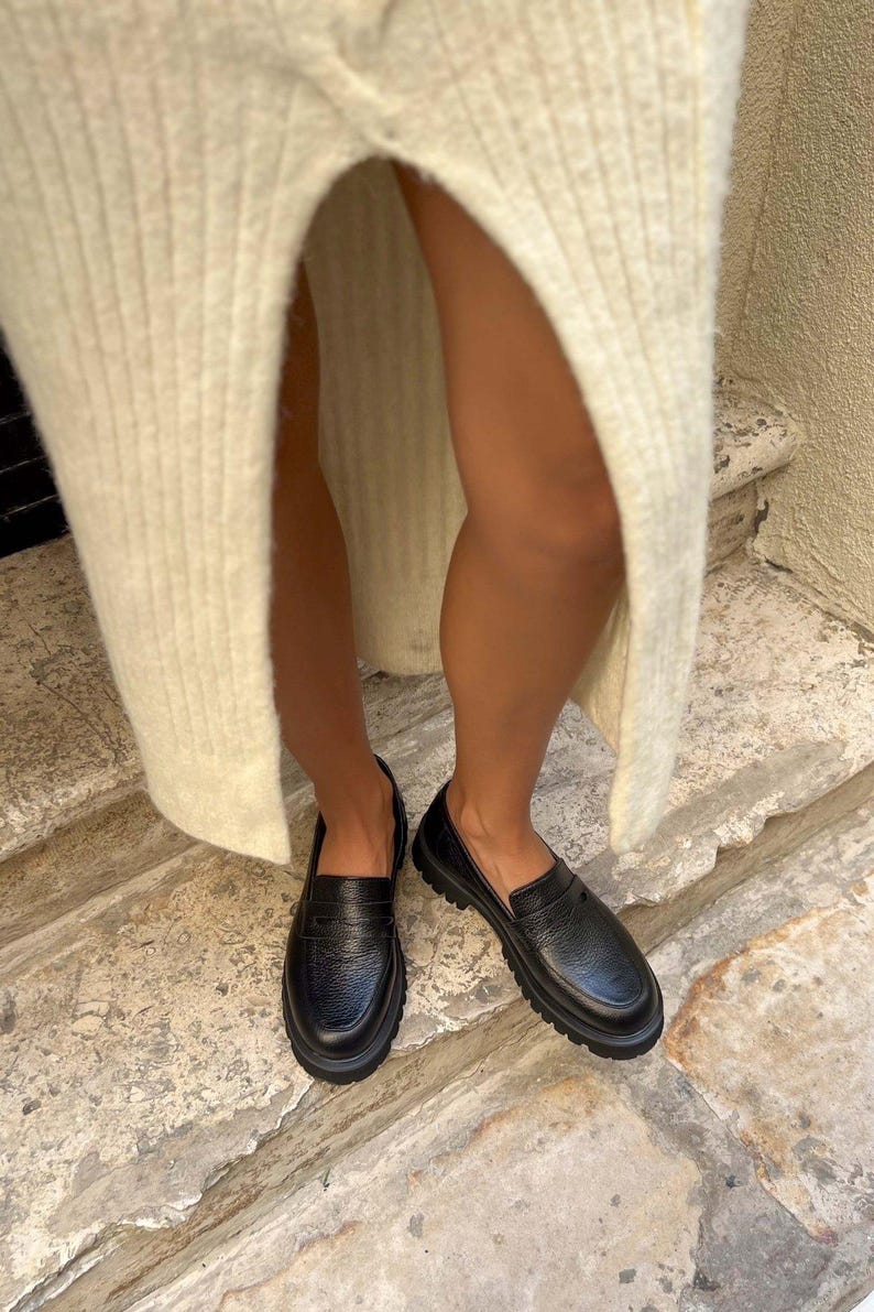 May include: Black leather loafers with a textured finish. The shoes have a chunky sole and a classic penny loafer design. The image shows the shoes from a low angle, highlighting their shape and texture. The loafers are worn with a cream-colored knit skirt.