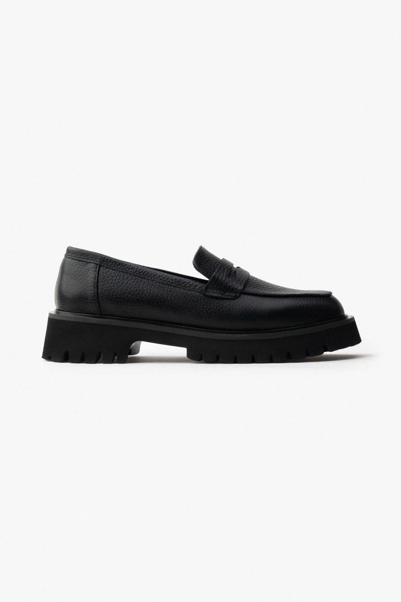 May include: Black leather loafer with a chunky, lug sole. The shoe features a classic penny loafer design with a textured leather upper and a low heel. The sole is thick and has a rugged tread for added grip. A stylish and comfortable shoe.