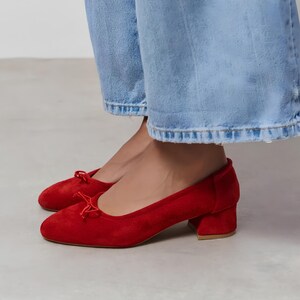 May include: A pair of bright red suede ballet flats featuring a rounded toe, a small block heel, and a delicate decorative bow detail on the front vamp. The shoes are worn with light blue denim jeans that have a frayed hemline at the bottom.