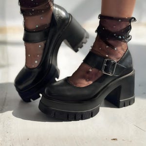 Può includere: Scarpe Mary Jane nere con plateau, tacco grosso e chiusura con fibbia. Le scarpe hanno una suola strutturata e una punta arrotondata. Sono abbinate a calzini neri trasparenti con un motivo a stelle.