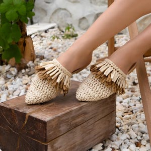 May include: Beige woven raffia mules with a fringe detail. The open-back slip-on shoes have a pointed toe. The fringe is a mix of light and dark brown. The shoes are resting on a wooden block.