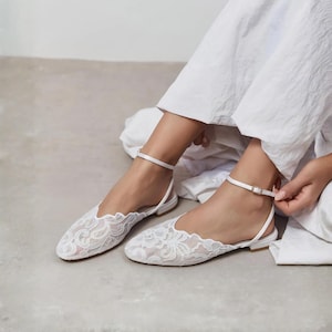 May include: White lace flats with ankle straps. The shoes feature a delicate floral lace pattern and a scalloped edge. The ankle straps have a small gold-colored buckle. The flats are designed for a formal occasion.