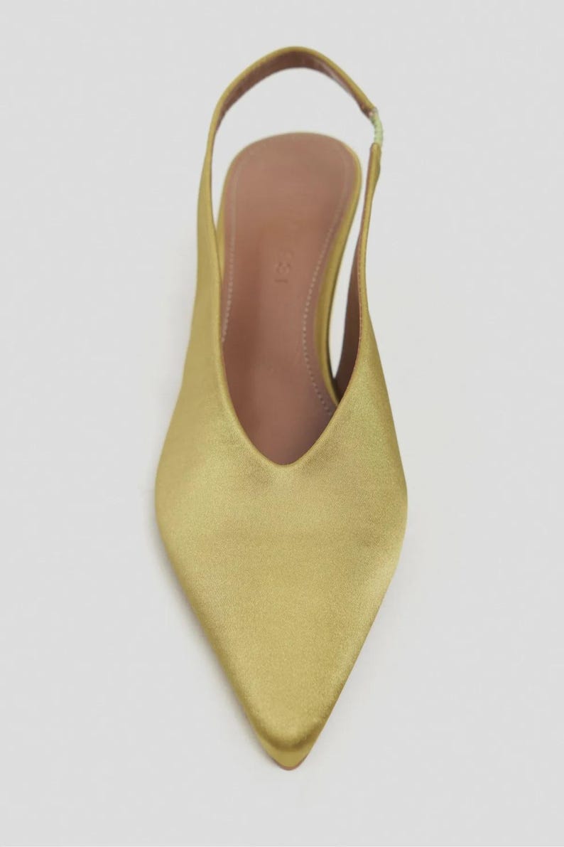 Może przedstawiać: But typu slingback ze szpiczastym noskiem w metalicznym, chartreuse satynowym wykończeniu. But posiada głębokie wycięcie w kształcie litery V, smukły obcas oraz dopasowany elastyczny pasek na pięcie. Wkładka wewnętrzna jest w neutralnym kolorze beżowym.