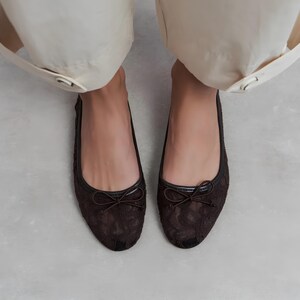 May include: A pair of dark brown ballet flats featuring a delicate lace overlay pattern across the entire upper. Each shoe is accented with a small, matching dark brown ribbon bow at the rounded toe and a smooth, dark trim along the collar.