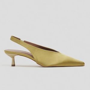 Może przedstawiać: Satynowy but typu slingback w kolorze chartreuse ze szpiczastym noskiem i niskim, metalicznym obcasem typu kaczuszka. But posiada cholewkę w kształcie litery V, cienki regulowany pasek na pięcie oraz gładkie, lśniące wykończenie odbijające światło.