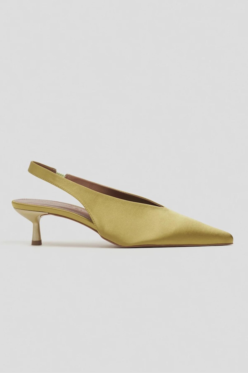 Może przedstawiać: Satynowy but typu slingback w kolorze chartreuse ze szpiczastym noskiem i niskim, metalicznym obcasem typu kaczuszka. But posiada cholewkę w kształcie litery V, cienki regulowany pasek na pięcie oraz gładkie, lśniące wykończenie odbijające światło.