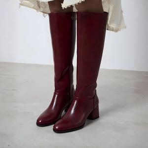 May include: A pair of knee-high burgundy leather boots with a block heel. The boots have a glossy finish and a zipper on the side. The boots are a solid color and have a classic design.