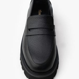 May include: Black leather loafer with a textured upper and a thick, lug sole. The shoe features a classic penny loafer design with a strap across the vamp. The sole is approximately 1.5 inches thick.