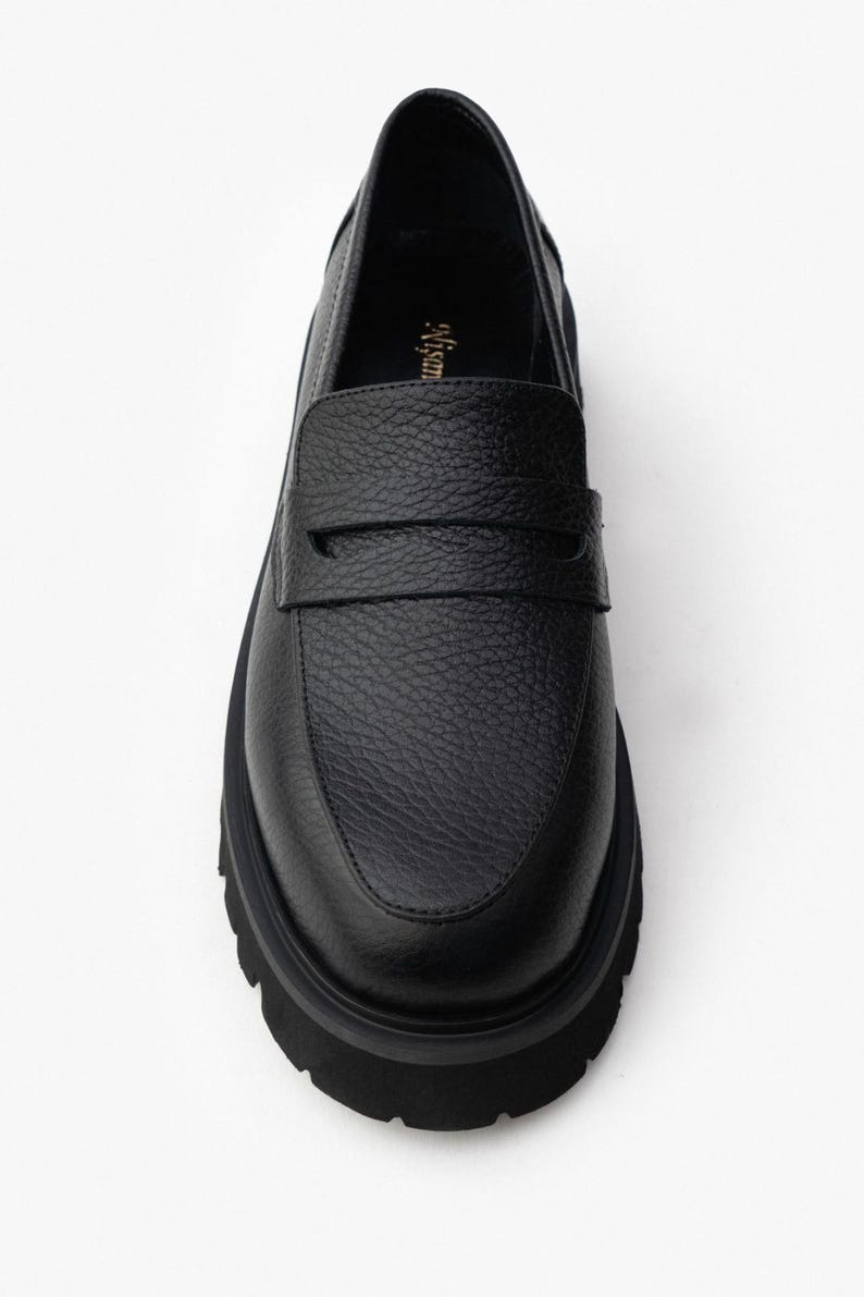 May include: Black leather loafer with a textured upper and a thick, lug sole. The shoe features a classic penny loafer design with a strap across the vamp. The sole is approximately 1.5 inches thick.