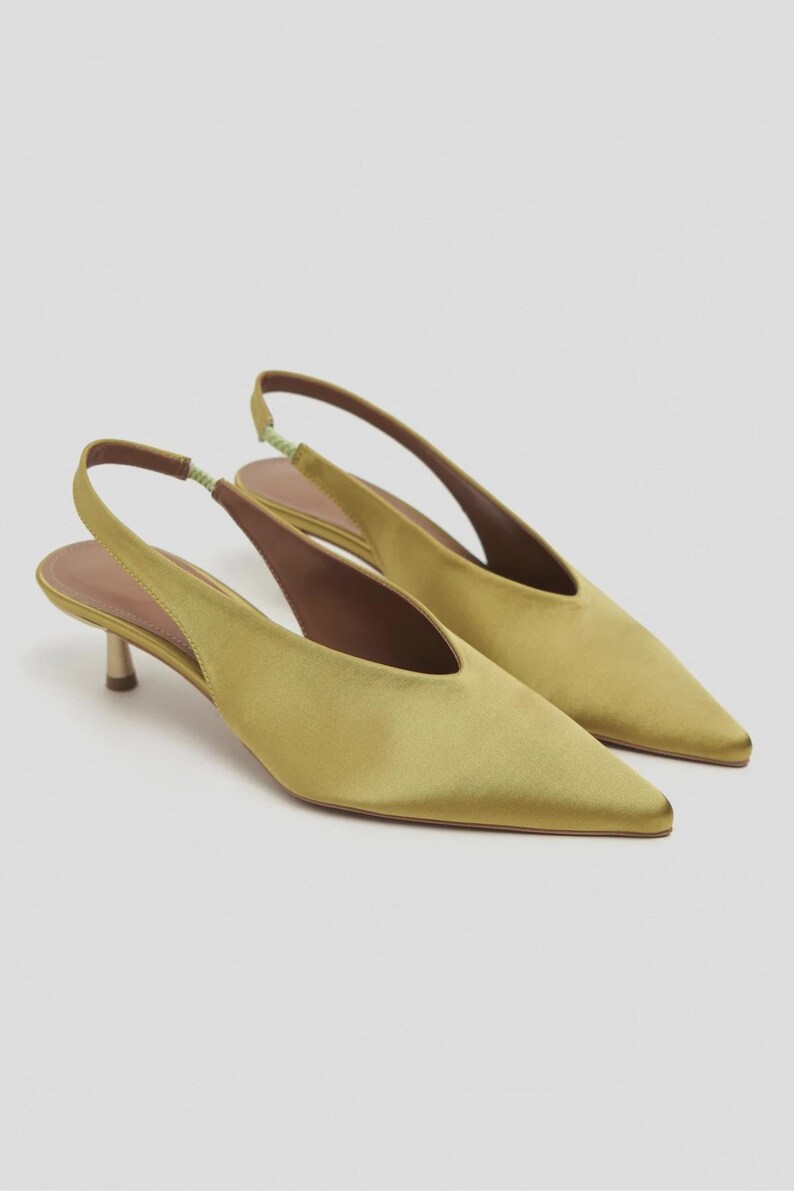Może przedstawiać: Para but&oacute;w typu slingback z satyny w kolorze chartreuse, ze szpiczastym noskiem, niskim obcasem typu kaczuszka oraz jasnozielonym elastycznym paskiem na pięcie. Wnętrze but&oacute;w wyłożone jest brązowym materiałem, o smukłym i prostym kroju.