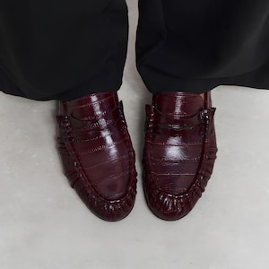 May include: A pair of dark burgundy loafers featuring a glossy, textured finish that mimics reptile skin. These slip-on shoes have a classic penny loafer strap across the top and a distinctive gathered, pleated edge detail around the rounded toe.
