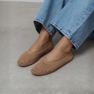 May include: A pair of tan crochet flats featuring a rounded toe and a woven, open-knit texture. The shoes have a low-profile silhouette with a simple slip-on design, showcasing a consistent tan yarn construction throughout the entire upper surface.