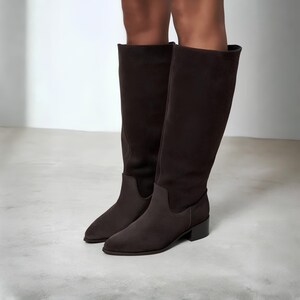 May include: A pair of dark brown knee-high boots with a smooth, matte finish. These boots feature a pointed toe, a structured shaft that reaches below the knee, and a low, block heel. The design is clean and simple with no visible hardware.