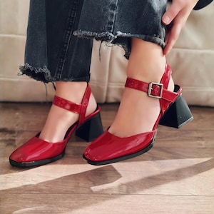 May include: Red patent leather Mary Jane style shoes with a square toe and a black geometric heel. The shoes have an ankle strap with a silver buckle. The shoes are on a wooden floor.