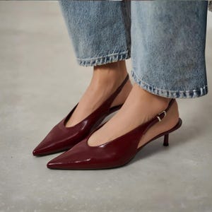 May include: Burgundy-colored slingback heels with pointed toes. The shoes have a glossy finish and a small, thin heel. A silver buckle secures the ankle strap. The shoes are worn with blue jeans.