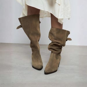 Puede incluir: Un par de botas altas de ante color topo con diseño de caña holgada, punta afilada y un pequeño detalle de hebilla lateral. Las botas tienen un tacón estructurado y una textura mate suave, extendiéndose hasta la pantorrilla con un ajuste relajado.