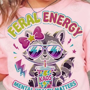 Peut inclure: Un t-shirt rose clair avec un motif de raton laveur portant des lunettes de soleil violettes et un nœud rose. Le raton laveur tient un gobelet marqué KAOS. Le texte indique FERAL ENERGY, MENTAL HEALTH MATTERS et YOU ARE NOT ALONE avec des éclairs.