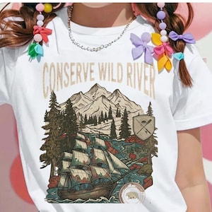 Può includere: T-shirt bianca con una grafica raffigurante una nave a vela su un fiume, montagne e la scritta "CONSERVE WILD RIVER". Il design include uno scudo con remi incrociati e un emblema di orso, nei toni del marrone, blu e verde.