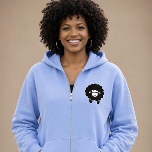 May include: A light blue zip-up hoodie featuring a black sheep graphic on the front. The hoodie has a drawstring hood and a full-length zipper. The text "BORN ALACYN" is printed vertically on the right sleeve.