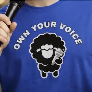 May include: A blue t-shirt with the text "OWN YOUR VOICE" in white, arched above a black sheep holding a microphone. The sheep graphic is in a cartoon style. The person is holding a black microphone.