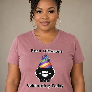 Op de afbeelding: Een mauve V-hals T-shirt met de tekst "Born Different" en "Celebrating Today". Het shirt heeft een afbeelding van een zwart schaap met een regenboogfeestmuts. De vrouw op de foto draagt gouden oorringen.