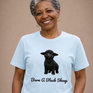 May include: Light blue t-shirt featuring a black sheep graphic and the text "Born A Black Sheep". The shirt is made of a soft material and has short sleeves. The graphic is a detailed illustration of a black sheep.