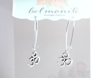 May include: Silver hoop earrings with a small Om symbol charm hanging from each hoop. The earrings are displayed on a card with the text "belmonili vintage reborn".
