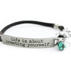 May include: A brown leather bracelet with a silver charm that reads "life is about creating yourself". The charm has a turquoise stone attached to it.