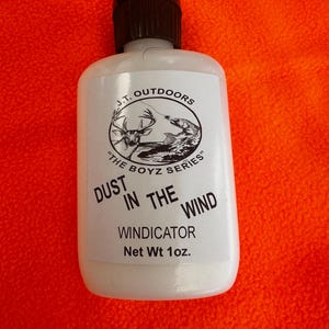 May include: A white bottle with a black cap, labeled "J.T. Outdoors" and "Dust in the Wind Windicator." The label features a deer and fish graphic. Net weight is 1 oz.