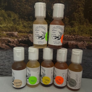 May include: A collection of small, clear bottles with white caps, labeled with various fishing scents. The labels feature text like "Garlic Scent," "Fish Oil Scent," and "Hatchery Blend." The bottles are arranged in a group, suggesting a product line for anglers.