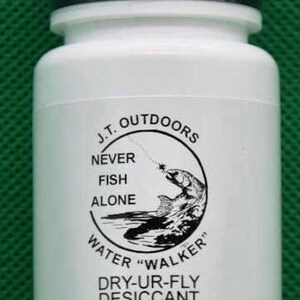May include: White plastic bottle with a black lid. The label features a circular graphic with a fish jumping out of the water and the text "J.T. OUTDOORS NEVER FISH ALONE WATER 'WALKER' DRY-UR-FLY DESICCANT Perfect for all flies".