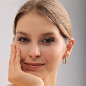 Op de afbeelding: Een vrouw draagt ​​gouden rechthoekige oorbellen met kleine, sprankelende steentjes. De oorbellen hebben een modern design, met een verticale staaf verbonden met een horizontale staaf. De vrouw heeft een lichte huid en lichtbruin haar.