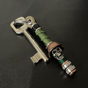 Könnte beinhalten: Ein dekorativer Schlüssel mit einem Totenkopf-Anhänger. Der Schlüssel hat eine silberne Oberfläche, und der Anhänger zeigt einen Totenkopf mit einem bronzefarbenen Helm. Der Anhänger ist mit grünem Kordel umwickelt und mit silbernen und kupferfarbenen Perlen akzentuiert.
