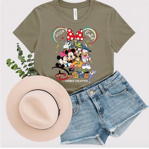 Puede incluir: Una camiseta verde oliva con un dise&ntilde;o de vacaciones familiares de Disney, con Mickey Mouse y sus amigos. La camiseta se combina con pantalones cortos de mezclilla y un sombrero rosa claro. El texto de la camiseta dice "2026 Emily".