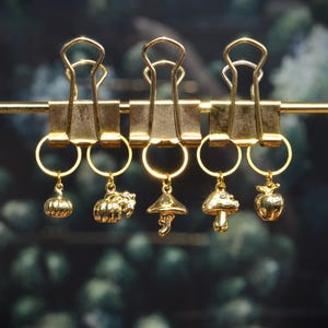 May include: Three gold-toned metal binder clips suspended on a horizontal gold rod. Attached to the clips are five gold-coloured charms featuring a small pumpkin, a larger pumpkin, a mushroom, a spotted mushroom, and a small apple shape.