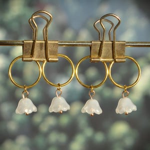 May include: Four gold-toned metal rings, each featuring a small, translucent white bell-shaped flower charm. The rings are suspended from two gold-toned metal binder clips attached to a thin horizontal metal rod, creating a uniform display.
