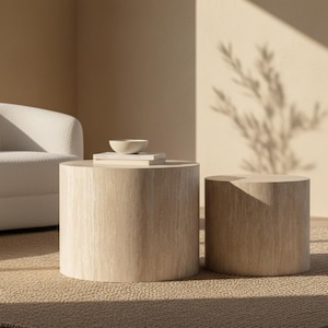 May include: Two cylindrical wooden coffee tables in a neutral-toned living room. The larger table holds a stack of books and a small white bowl. The room features a white sofa and a textured rug, with sunlight casting shadows.