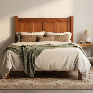 May include: A wooden bed frame with a tall, paneled headboard featuring a light-colored duvet, two beige pillows, two brown rectangular pillows, and a sage green knitted throw blanket draped across the center of the bed.
