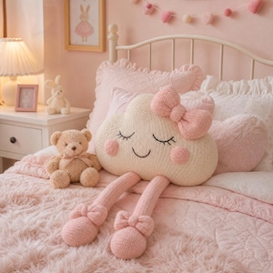 Puede incluir: Un cojín de peluche con forma de nube de color crema y textura tejida, que presenta una cara sonriente con ojos cerrados, mejillas rosadas y un lazo rosa. El cojín tiene dos piernas largas y colgantes tejidas con lazos rosas en los tobillos.