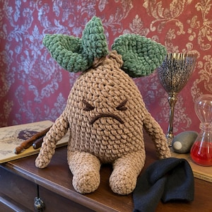 Peut inclure: Une peluche crochetée en forme de racine de mandragore avec un corps texturé marron clair et deux grandes touffes de feuilles vert foncé sur le dessus. Le jouet présente un visage brodé à l'expression grincheuse, des yeux fermés et deux bras.
