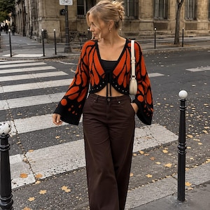 Könnte beinhalten: Eine bauchfreie, schwarz-orangefarbene Strickjacke mit Schmetterlingsflügelmuster, langen ausgestellten Ärmeln und Bindeband vorne, kombiniert mit einem schwarzen Oberteil, dunkelbrauner weiter Hose, einer kleinen cremefarbenen Umhängetasche und beigen Plateauschuhen.