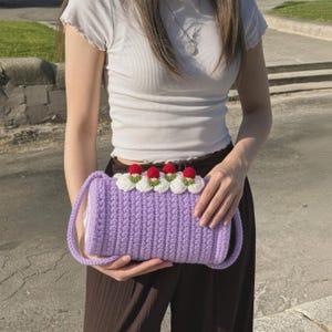May include: A cylindrical crochet handbag in a light purple shade, featuring a textured stitch pattern. The top edge is decorated with four white crocheted flowers, each topped with a small red berry accent, and includes a matching purple strap.