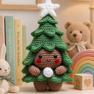 Op de afbeelding: Een gehaakte pop in de vorm van een kerstboom met een bruin gezicht, ronde zwarte ogen en rode wangen. De pop draagt een groen gelaagd boomkostuum met een witte ster op de top en houdt een kleine witte pompon vast in de gehaakte bruine handen.