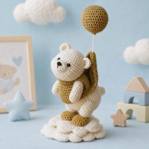 Puede incluir: Una figura de oso tejida a ganchillo de color crema con un chaleco amarillo mostaza, de pie sobre una base de nube blanca tejida a ganchillo. El oso sostiene un hilo blanco fino unido a un globo redondo amarillo mostaza sobre su cabeza.