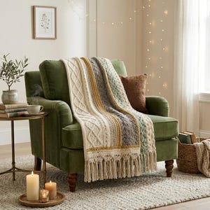 Pode incluir: Uma manta de malha grossa com painéis verticais em creme, amarelo mostarda, cinza e verde sálvia, com padrões de tranças intrincados e franjas longas, drapeada sobre uma poltrona de veludo verde-oliva escuro com uma almofada castanha.