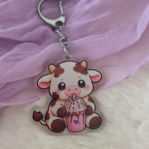 Könnte beinhalten: Ein Schlüsselanhänger aus klarem Acryl mit einer Cartoon-Kuh mit braunen Flecken, die sitzt und ein rosa Getränk mit einem violetten Herzsymbol auf dem Becher hält. Die Kuh hat große schwarze Augen und trinkt durch einen Strohhalm.